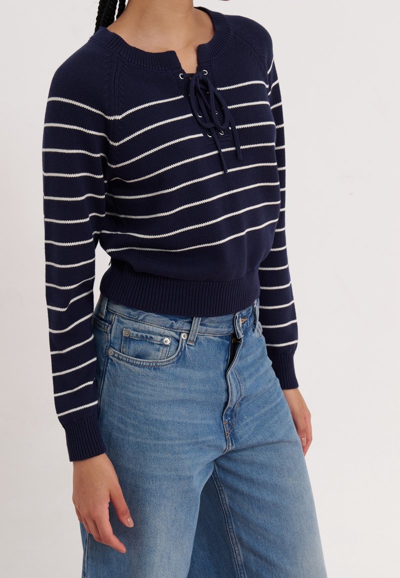 Femme portant un pull bleu marine avec des rayures horizontales blanches et un détail noué au niveau du col, associé à un jean bleu taille haute.