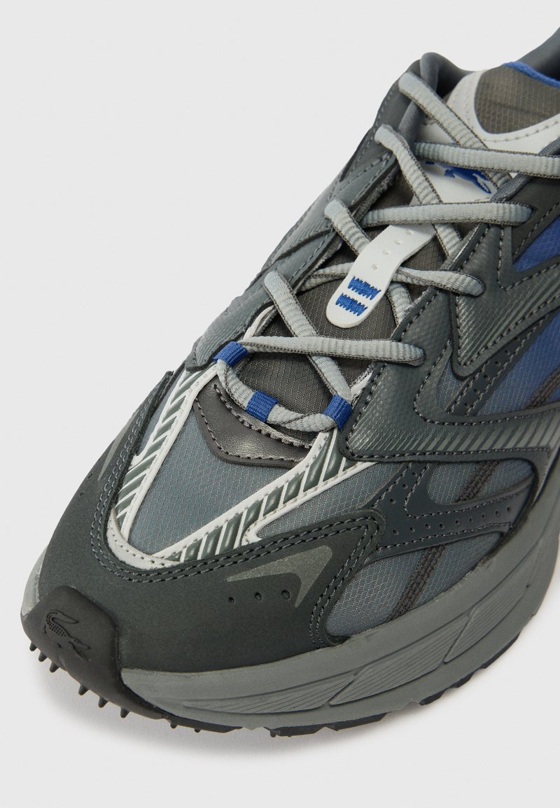 Nærbillede af en grå og blå atletisk sneakers med detaljeret syning, krydsede snørebånd og en tekstureret sål på en hvid baggrund.