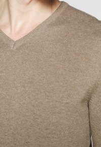 Beige V-ringad tröja gjord av stickat tyg, med en slät textur och färdiga kanter, med en tät passform vid halsen och axlarna.