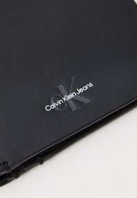 Svart texturerad material med präglad Calvin Klein Jeans-logotyp, med en kontrasterande grå "CK" designdetalj. Vikta kanter med sömnad.