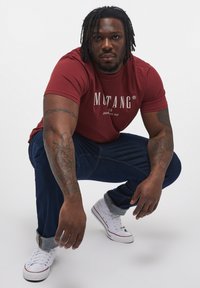Tricou roșu cu mânecă scurtă și logo alb, blugi negri, și adidași albi cu manșete gri. Modelul este în genunchi, evidențiind tatuajele de pe brațe.
