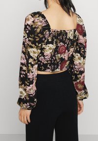 Blusa floreale cropped con sfondo nero, caratterizzata da fiori rosa, bianchi e bordeaux. Maniche lunghe con polsini elastici e scollatura quadrata.