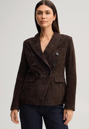 Vrouw draagt een donkerbruine double-breasted suède blazer met zwarte knopen en donkerblauwe broek tegen een effen achtergrond.