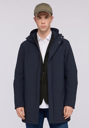 Parka blu navy con cappuccio, cerniera frontale e bottoni a pressione. Indossato sopra un gilet scuro e una camicia bianca. Cappello beige per un tocco di contrasto.
