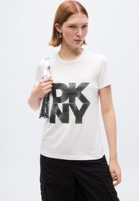 Λευκό βαμβακερό μπλουζάκι με μεγάλο μαύρο γκλίτερ λογότυπο "DKNY". Το μοντέλο κρατάει μια λαμπερή ασημένια τσάντα. Μαύροι παντελόνια cargo φαίνονται από κάτω.