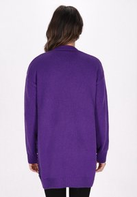 Cardigan viola lungo e sbagliato realizzato in materiale a maglia. Presenta polsini e orlo a coste, con dettagli decorativi lungo i lati.