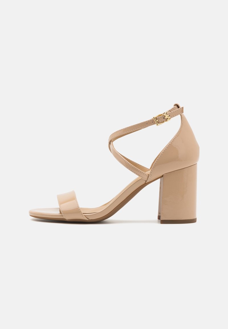 Sandale à talon en cuir verni beige avec un talon carré, une lanière unique sur les orteils et une lanière de cheville ajustable avec des accessoires dorés.