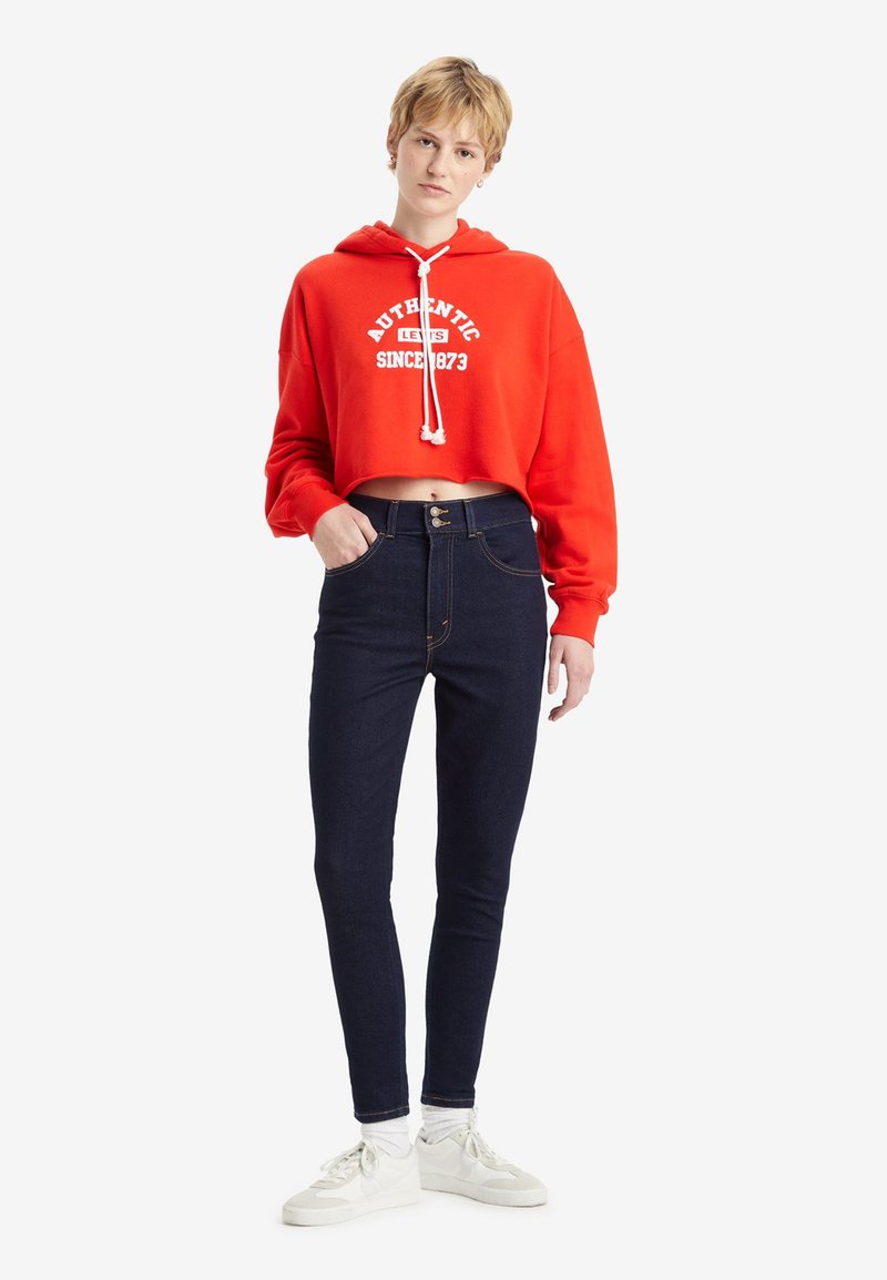Un hoodie rouge court avec un texte graphique blanc, associé à un jean taille haute bleu foncé et des baskets blanches, debout sur un fond uni.