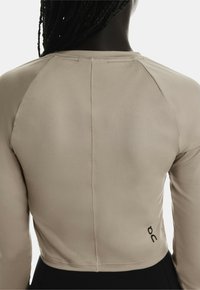 Haut court à manches longues en beige avec une texture lisse, présentant un détail de couture dans le dos et un petit logo sur le côté.