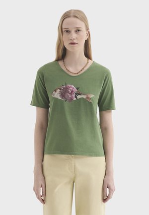 Frau mit langen blonden Haaren, die ein grünes T-Shirt trägt, auf dem ein Fisch mit einer rosa Blume auf dem Körper abgebildet ist, und hellbeige Hosen.