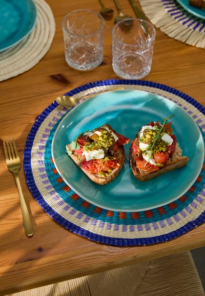 Deux tranches de pain grillé garnies de tomates colorées mélangées, de fromage de chèvre et de noix vertes, servies sur une assiette turquoise avec un dessous de plat tressé à motifs.