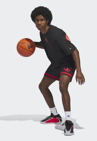Young man in black and red basketball outfit dribbling basketball, wearing white socks and black-red sneakers on plain background.