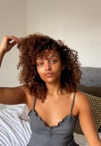 Harmaa ribbikuvioitu hihaton toppi aaltoilevilla reunoilla, rento istuvuus. Taustalla on sänky, jossa on tekstuurillisia tyynyjä ja peittoja.