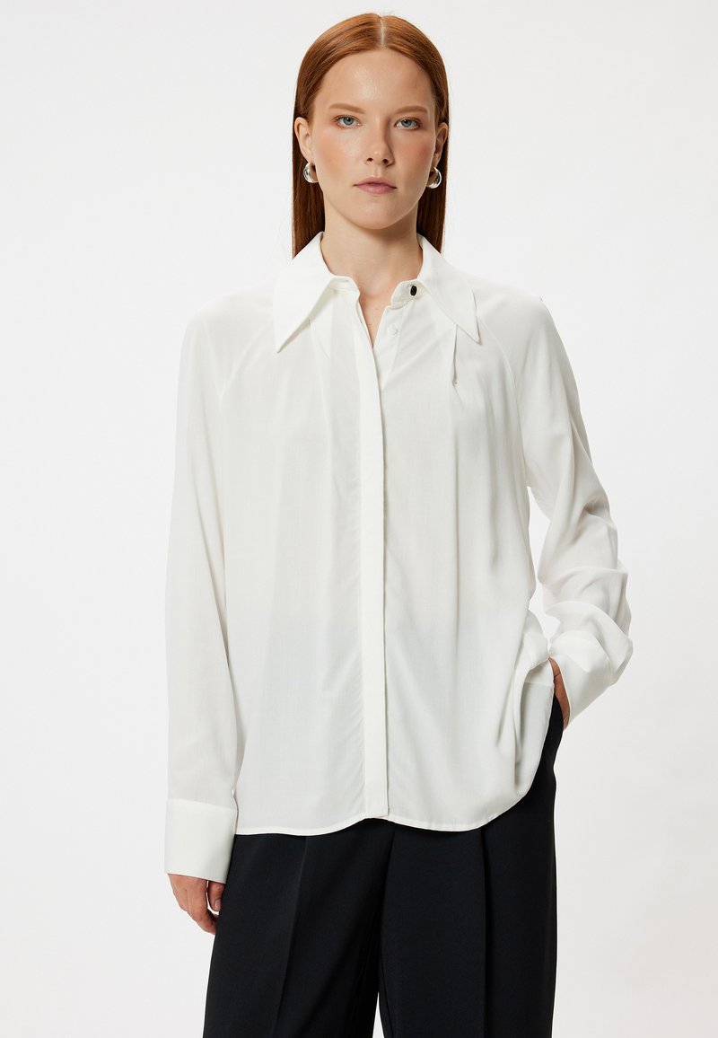 Blusa blanca de manga larga con cuello puntiagudo, botones frontales y un corte relajado. Tejido suave con un ligero brillo. Combinada con pantalones negros.