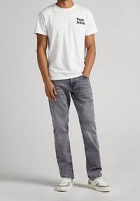 Camiseta blanca de algodón con el logo negro de "Pepe Jeans", combinada con jeans de mezclilla de color gris claro y zapatillas grises, de pie sobre un fondo neutro.