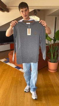 Siva majica z dolgimi rokavi in ovratnikom, vsebuje majhen vezenin logotip, etiketa še vedno pritrjena, drži jo oseba v športnih kavbojkah in supergah.