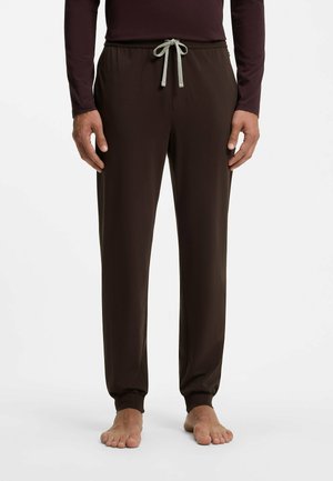 Man die donkerbruine loungebroek met een lichtgrijze koord draagt, gecombineerd met een donker topje met lange mouwen, staande op blote voeten voor een witte achtergrond.