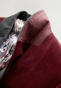 Burgundi bársony zakó sima külsővel, fekete díszítéssel és virágmintás belső béléssel, amely lila és fehér árnyalatú.