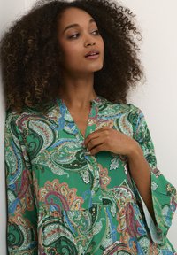 Zelena bluzica s paisley vzorom z V-izrezom, nabranim pasom in širokimi rokavi. Tkanina je okrašena z večbarvnimi cvetličnimi in paisley motivi.