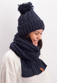 Dark navy knitted beanie with a pom-pom and scarf, featuring ribbed texture and leather accent. Cozy and thick, ideal for cold weather.