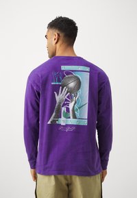 Maglietta a maniche lunghe viola con un design grafico di mani che tengono un pallone da basket, con accenti pastello e il testo "NIKE". Materiale morbido e casual.