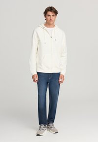 Giovane uomo in piedi dritto, vestito con un felpa bianca con zip, jeans blu e sneaker grigie, su uno sfondo grigio chiaro uniforme.