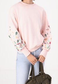Donna che indossa una felpa leggera color rosa con motivi floreali sulle maniche, jeans azzurri chiari, mentre tiene una borsa in corduroy verde per le maniglie.
