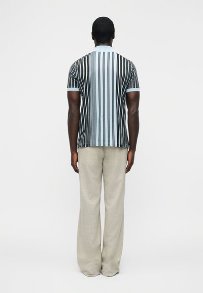 Άνδρας όρθιος με την πλάτη στην κάμερα, φορώντας κοντομάνικη, κάθετα ριγέ πόλο μπλούζα και μπεζ φαρδιά παντελόνια, μπροστά σε απλό φόντο.
