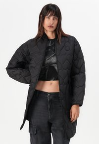 Zwarte gewatteerde jas met een hoge kraag en geribde manchetten, voorzien van een ritssluiting aan de voorkant en een rond logo-detail, gecombineerd met een zwarte cropped top en donkere jeans.