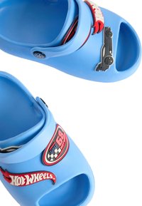 Blå plastträsko med upphöjda gummilogotyper från Hot Wheels, öppen tå-design och utskärningar på sidorna för ventilation. Platta sulor för komfort.