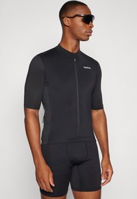 Mies pukeutunut mustaan lyhythihaiseen pyöräilypaitaan ja vastaaviin shortseihin, mustat wraparound-aurinkolasit päässään, seisoo yksivärisen taustan edessä.