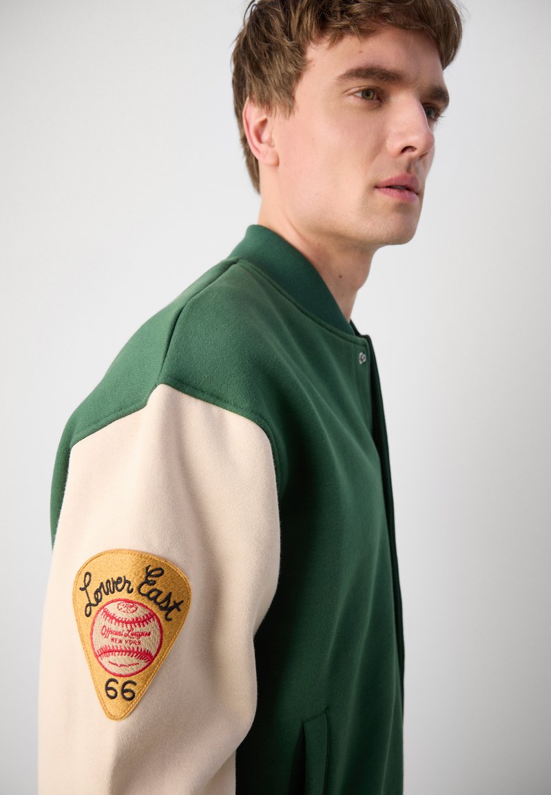 Veste universitaire verte avec des manches crème, arborant un patch doré avec "Lower East" et un design de baseball. Le matériau est doux et texturé.