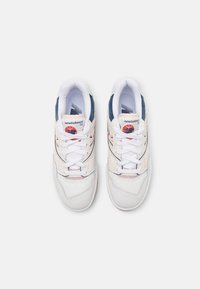 White and light pink sneakers with navy accents, featuring a textured upper, round toe, and white laces. Branding visible on tongue.