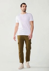 Mann iført hvit kortermet henley-skjorte, olivengrønne cargo-bukser og beige sneakers som står mot en lys bakgrunn.