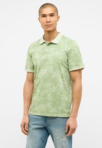 Hellgrünes Poloshirt mit einem strukturierten, blättrigen Muster, mit Kragen und einer Knopfleiste mit drei Knöpfen, kombiniert mit ausgewaschenen blauen Jeans.