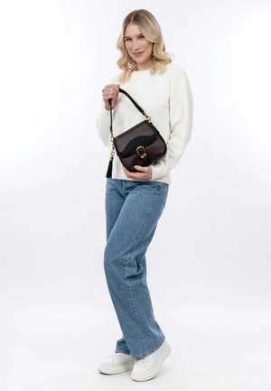 Borsa a tracolla in pelle marrone con dettagli neri, chiusura a fibbia e dettaglio con frange. Abbinata a un maglione bianco e jeans azzurri.