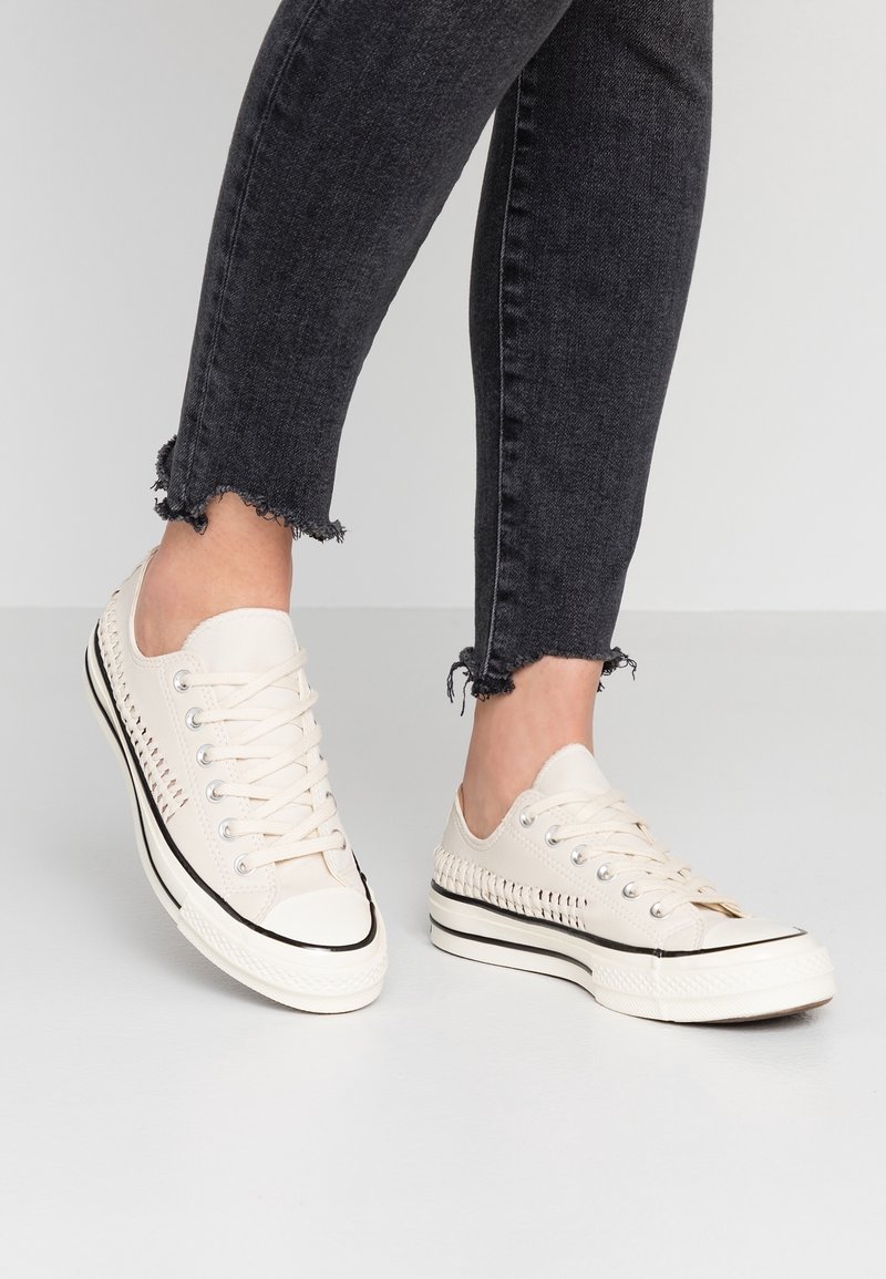 Baskets en toile beige avec un design latéral tissé texturé, semelle en caoutchouc blanche et bande d'accent en caoutchouc noir. Portées avec un jean noir effiloché.