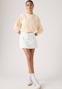 Γυναίκα με τα μαλλιά πιασμένα πίσω, φοράει χρυσούς κρίκους για σκουλαρίκια, μπεζ φούτερ με μακριά μανίκια, λευκή φούστα με κουμπιά μπροστά, λευκές κάλτσες και αθλητικά παπούτσια.