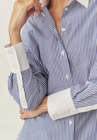 Camicia a righe blu e bianche con colletto classico, polsini bianchi e due bottoni su ogni polsino. Il tessuto è liscio e lucido.