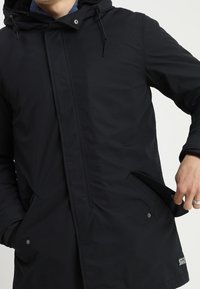 Veste noire imperméable avec capuche, dotée d'une fermeture à pression à l'avant, de poches latérales et d'un tissu texturé, conçue pour la fonctionnalité et la chaleur.