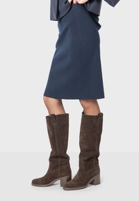 Bottes marron en suède, jusqu'au genou, avec un talon carré, assorties à une jupe crayon bleu marine, présentant une texture lisse et une silhouette ajustée.