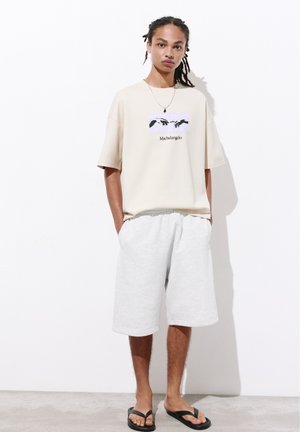 Jeune personne aux cheveux tressés porte un t-shirt beige Michelangelo, un short blanc au genou et des tongs noires, debout contre un mur blanc.