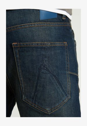 Tamsiai mėlynos džinsinės kelnės su oranžinėmis siūlėmis, išskirtiniu kišenių dizainu ir mėlynu odiniu logotipu ant juosmens.