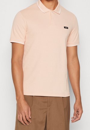 Hellrosa Poloshirt mit weißem Kragen und Ärmelsaum, mit kleinem schwarzem Logoaufnäher auf der linken Brust. Kurze Ärmel und klassischer Schnitt.