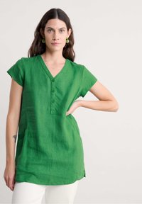 Tunika in lino verde con scollo a V, maniche corte e dettagli con bottoni. Presenta spacchi laterali e un tessuto strutturato, indossata con pantaloni bianchi.