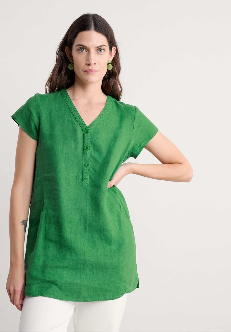 Tunika in lino verde con scollo a V, maniche corte e dettagli con bottoni. Presenta spacchi laterali e un tessuto strutturato, indossata con pantaloni bianchi.