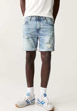 Homme portant un short en jean bleu clair déchiré, des chaussettes blanches, des baskets blanches et bleues, et un t-shirt blanc sur un fond uni.