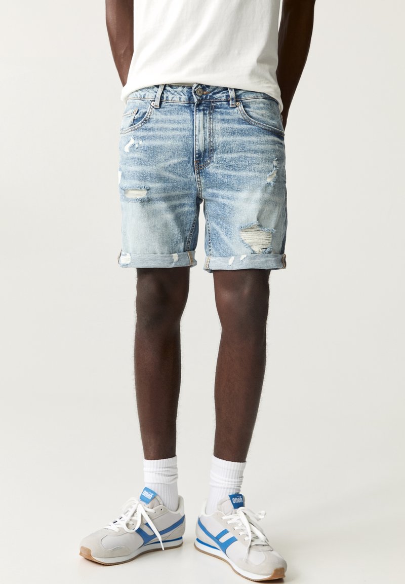 Homme portant un short en jean bleu clair déchiré, des chaussettes blanches, des baskets blanches et bleues, et un t-shirt blanc sur un fond uni.