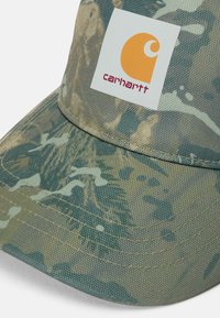 Gorra de camuflaje verde con visera plana, tejido texturizado y un parche cuadrado blanco con un logotipo naranja y el texto "carhartt".