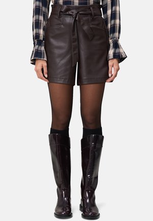 Shorts in pelle marrone con una cintura e tasche laterali, indossati con stivali neri alti fino al ginocchio in vernice e collant neri trasparenti.